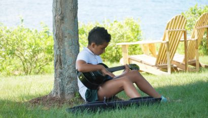 A camper sitting under a tree playing the guitar.