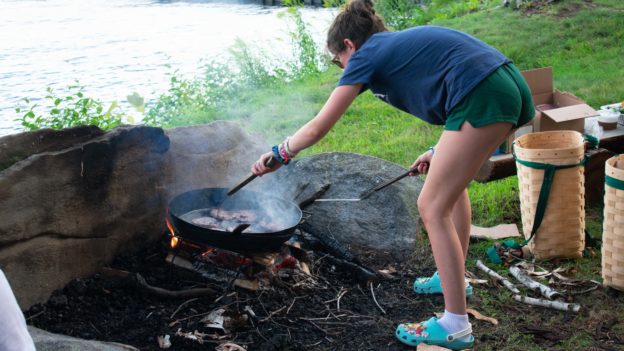 A camper cooking over an open flame by a lake.
