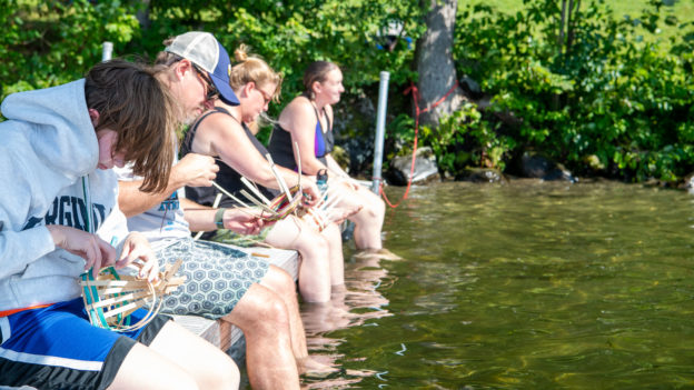 Campers weaving baskets while dipping their feet in a lake.