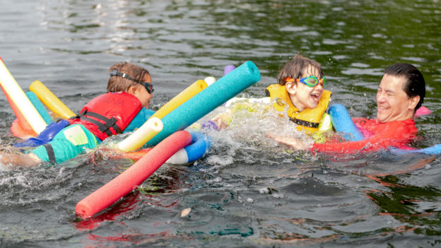 Campers swimming in a lake with colorful pool noodles.