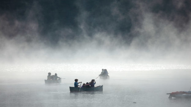 A lake shrouded in mist with people on canoes.