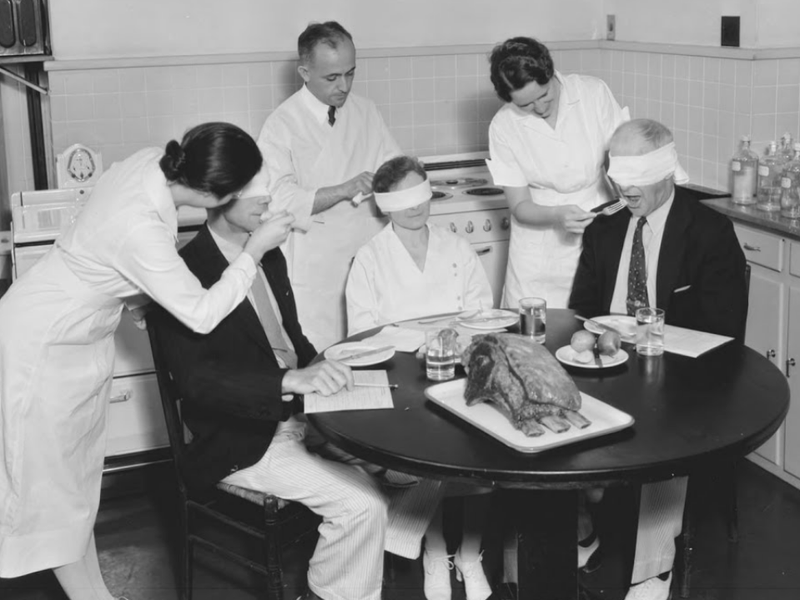 A black and white photo of a group eating a meal blindfolded.