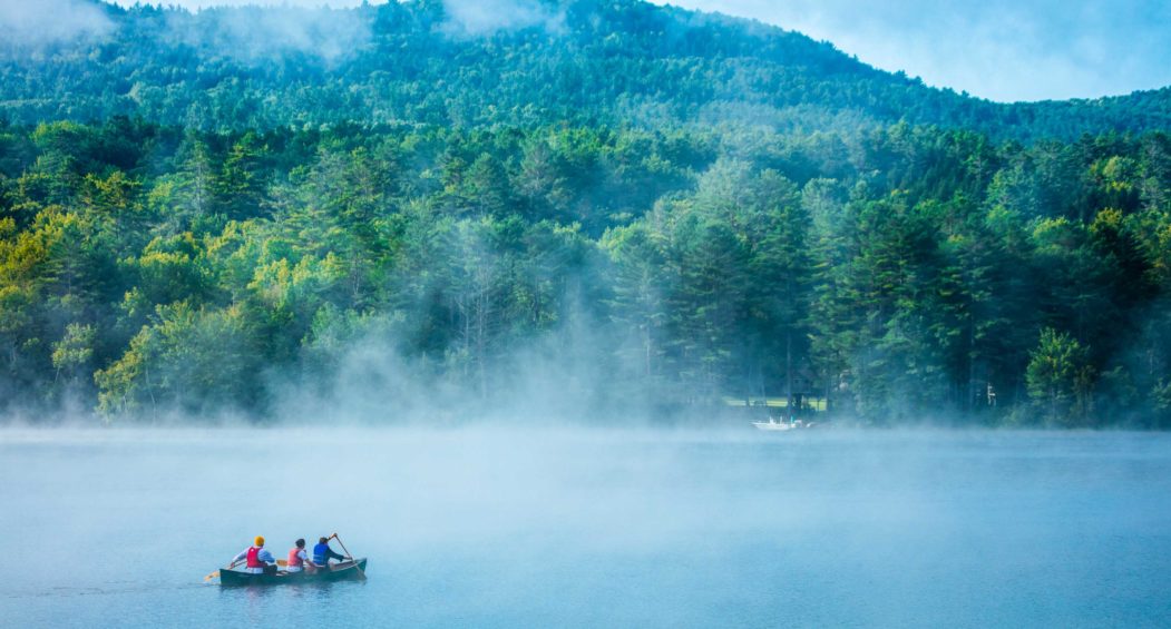 A lake shrouded in mist along with three people canoeing.