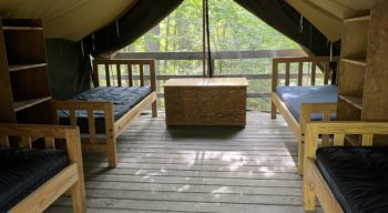 Interior of a canvas tent with wooden floors, featuring four wooden beds with black mattresses, a central wooden storage chest, and shelves.