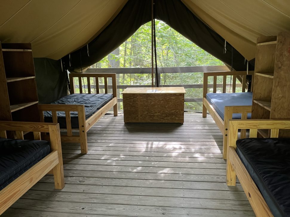 Interior of a canvas tent with wooden floors, featuring four wooden beds with black mattresses, a central wooden storage chest, and shelves.